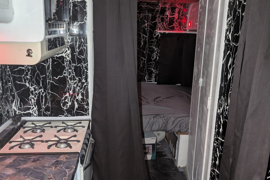 Inside an RV rental: a stylish black and white marble kitchen area with a stove, and a glimpse of a compact sleeping space.