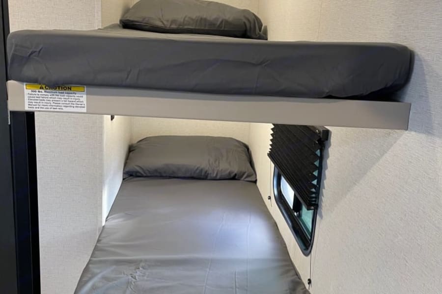Inside an RV rental, two bunk beds with grey bedding offer compact sleeping. Note the top bunk's weight limit label and window below.