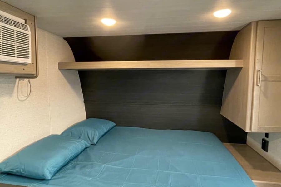 Comfortable RV rental bedroom: blue sheets on bed, overhead shelf, AC unit, and built-in storage for travel.