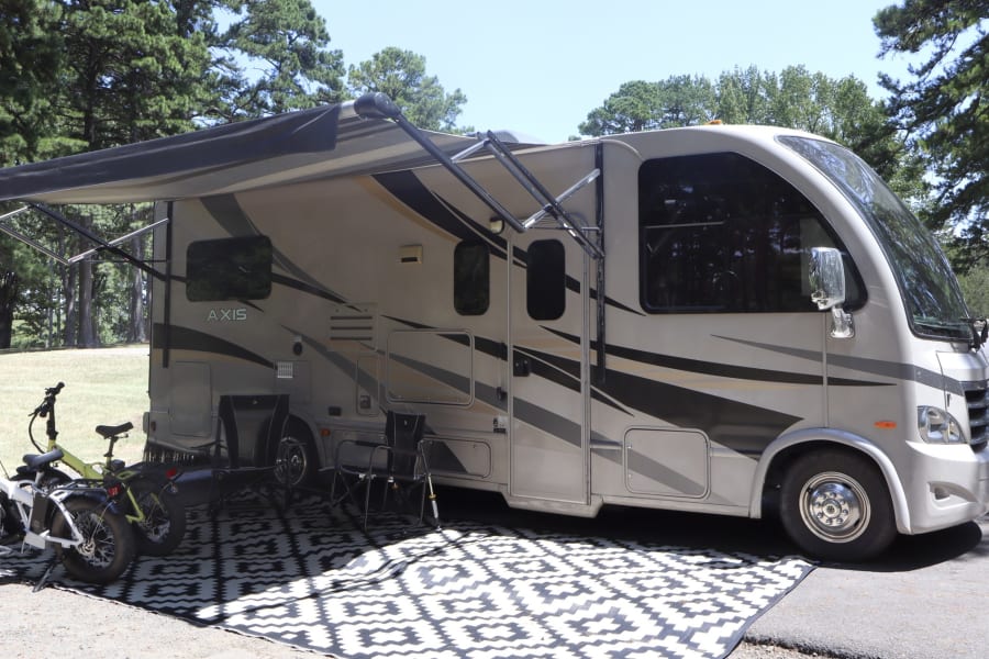 A beige Thor Axis RV rental with an extended awning provides shade over e-bikes and an outdoor mat at a sunny campsite.
