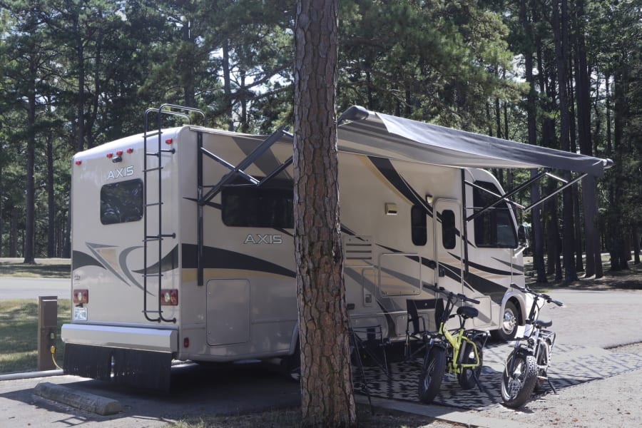 Spacious RV rental parked at a serene campsite with an awning out and two e-bikes ready for adventure among pine trees.