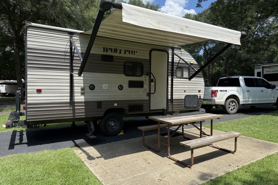 Enjoying the outdoors with an RV rental; Wolf Pup travel trailer, awning, & picnic table at a shaded campsite.