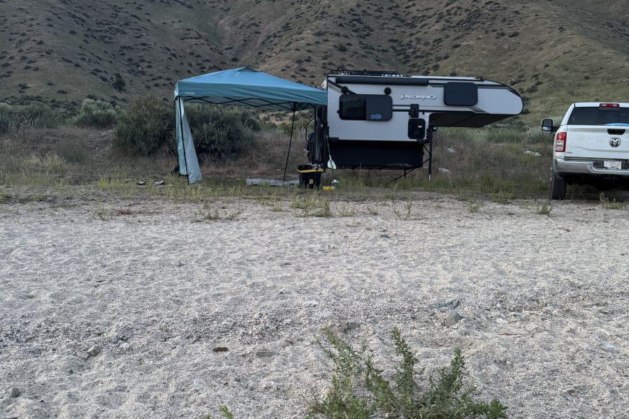 A grey truck with an RV rental truck camper & a blue canopy set up at a gravel campsite, backed by vegetated hills.