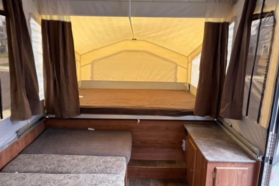 Inside a pop-up RV rental: a bed with brown mattress, patterned bench seating, and wood cabinetry on the right.