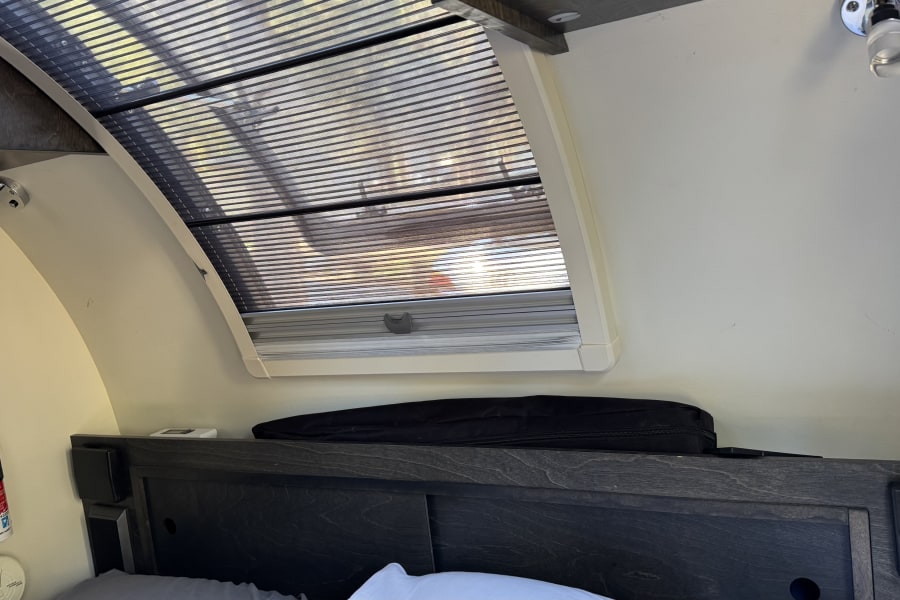 Cozy RV rental bedroom featuring a skylight with blinds, a reading light, and a mounted fire extinguisher for safety.
