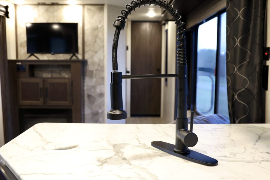 This rv rental features a modern kitchen with a sleek black faucet, marble-look countertop, and a cozy living area with TV.