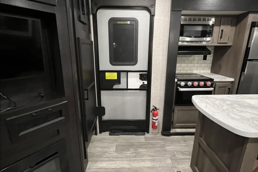 RV rental interior: living area with dark cabinetry, main entry door with fire extinguisher, and kitchen with stove, microwave, and island.