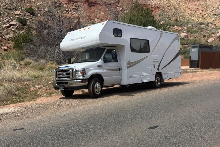 A white Minnie Winnie RV rental parked on a desert road shoulder, with stunning red rock mountains as the backdrop.