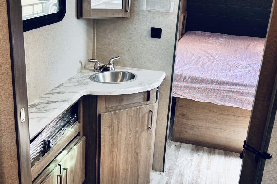 Inside an RV rental: a compact living area featuring a light wood vanity with a stainless steel sink, a mirror, and a cozy bedroom beyond.