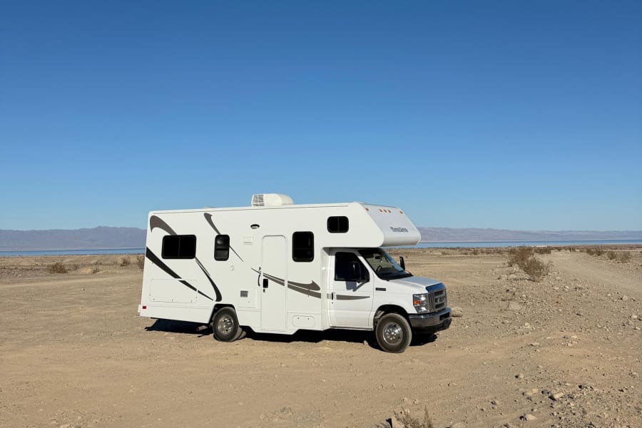 A white RV rental is parked on a dirt road in a desert landscape with distant mountains, water, and a clear blue sky, ideal for travel.