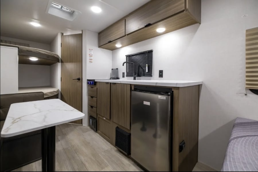 Explore this modern RV rental interior, showcasing a compact kitchen with a fridge, sink, and ample cabinetry, plus bunk beds and a dining area.
