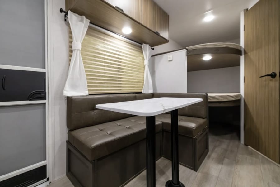 Cozy RV rental interior: L-shaped dinette, window with blinds, overhead storage, and bunk beds in back.