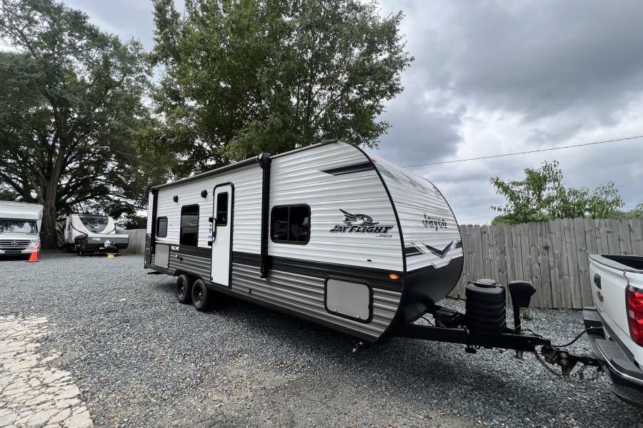 White Jayco Jay Flight travel trailer for RV rental, connected to a pickup truck in a gravel lot with other RVs under cloudy skies.