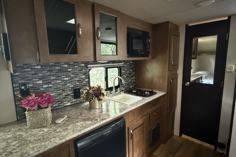 Spacious RV rental kitchen with wood cabinets, stone-look counters, sink, stove, microwave, and a private bedroom view.
