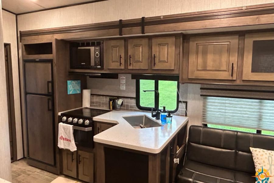 This RV rental interior shows a full kitchen with microwave, stove, sink, and fridge, plus a cozy sofa. Ready for adventures!
