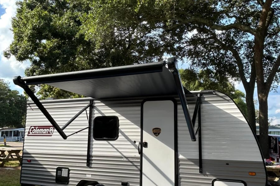 Coleman RV rental at a sunny campsite with an extended awning, chairs, and picnic table under large trees.