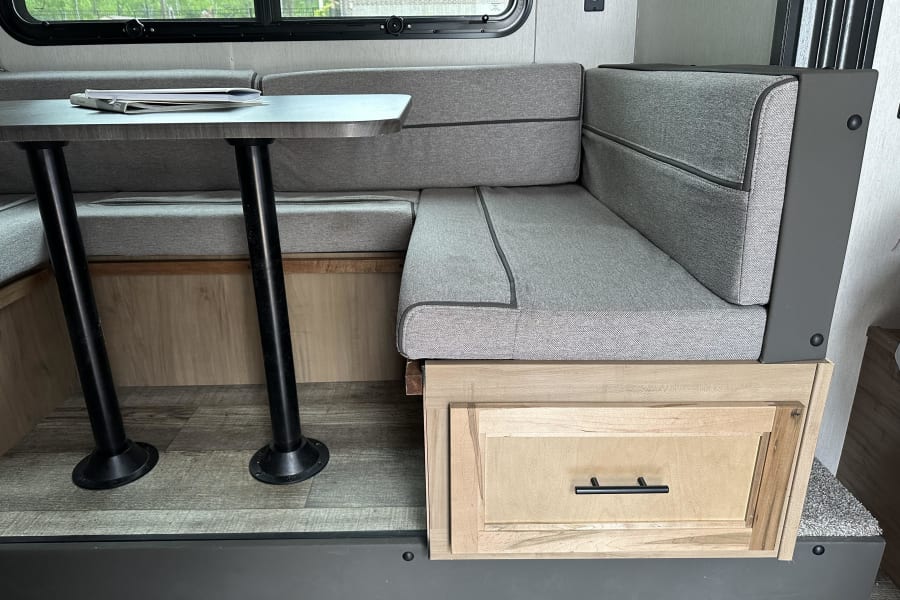 Modern RV rental interior: Dinette booth with gray fabric cushions, table, window, and wooden storage drawer.