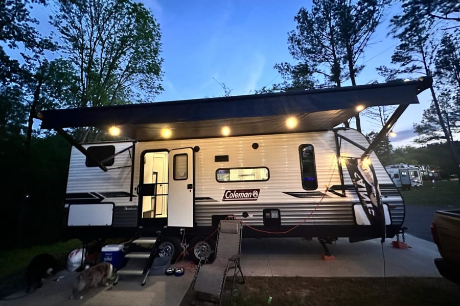 Enjoying a peaceful evening at a campsite with an RV rental. This travel trailer has an illuminated awning and open door, with dogs nearby.