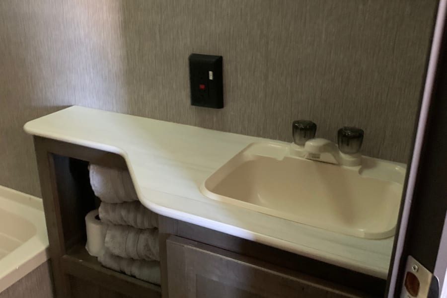 RV rental bathroom: compact vanity with sink, mirror, and handy towel storage. Toilet & tub partly visible.