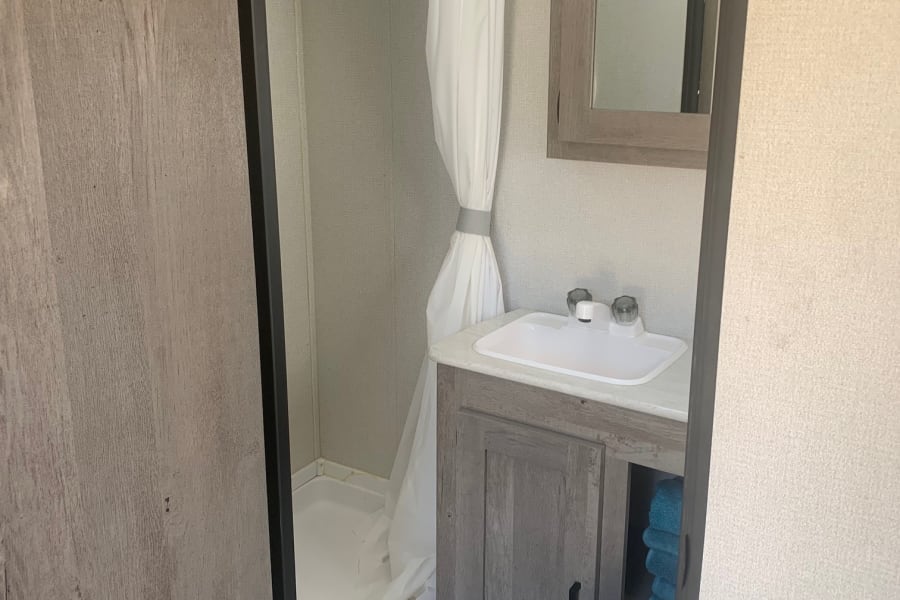 Compact RV rental bathroom with shower, sink, mirrored vanity, toilet, and blue towels. Clean and functional.