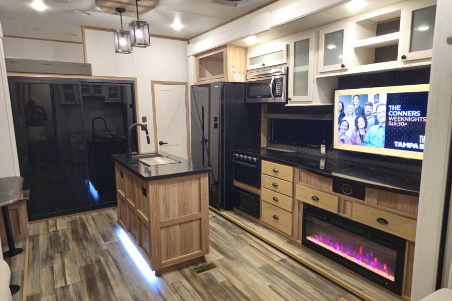 An RV rental's spacious interior with a modern kitchen island, black appliances, a large TV, and an electric fireplace.