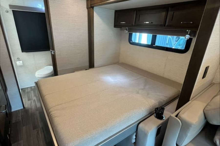This RV rental interior features a fold-down bed with a sofa, overhead cabinets, and a bathroom entrance. Efficient compact living.