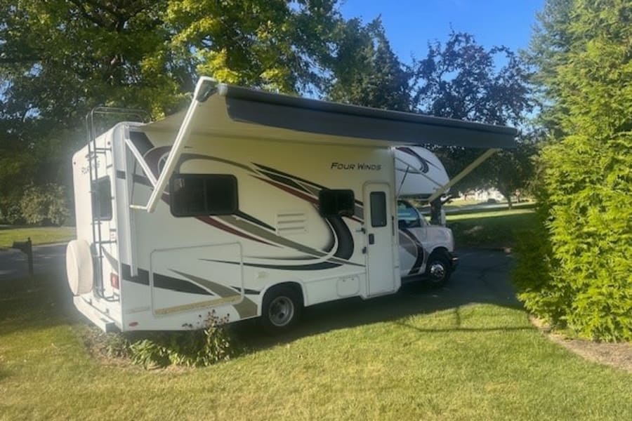 White Four Winds motorhome rv rental parked on green grass with awning out, surrounded by trees.