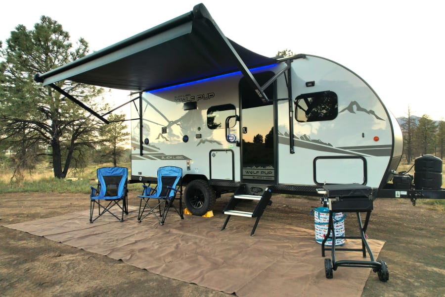RV rental comfort! A Wolf Pup Black Label travel trailer with an illuminated awning, chairs, and grill set up for camping.