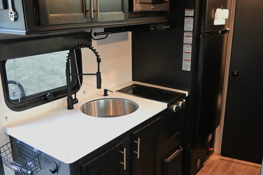 Explore this stylish RV rental kitchen: black cabinets, white countertop, sink, stove, microwave, and fridge. Ready for your journey.
