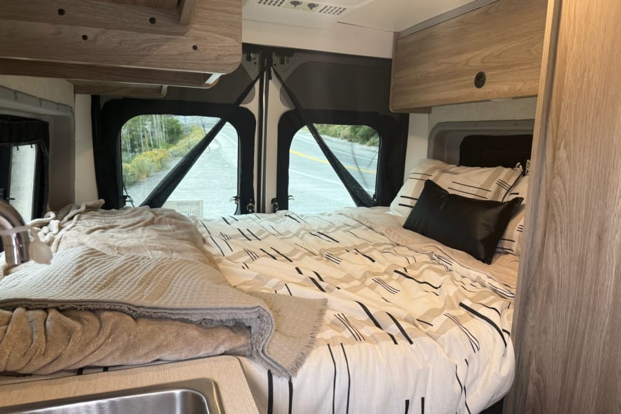 Inside a compact rv rental: cozy bed with graphic bedding, small sink, overhead cabinets, and a scenic view through rear windows.