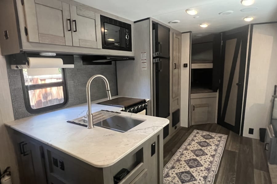 Modern RV rental kitchen: sink, counter, microwave, fridge, bunk beds & floor rug, lit by overhead lights.