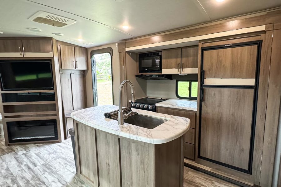 Spacious, modern rv rental interior featuring a full kitchen island, stove, fridge, entertainment center, and fireplace.