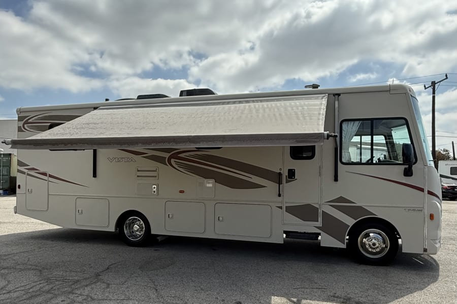 A beige Winnebago Vista RV rental with an extended awning is parked on pavement under a cloudy sky. Perfect for travel!