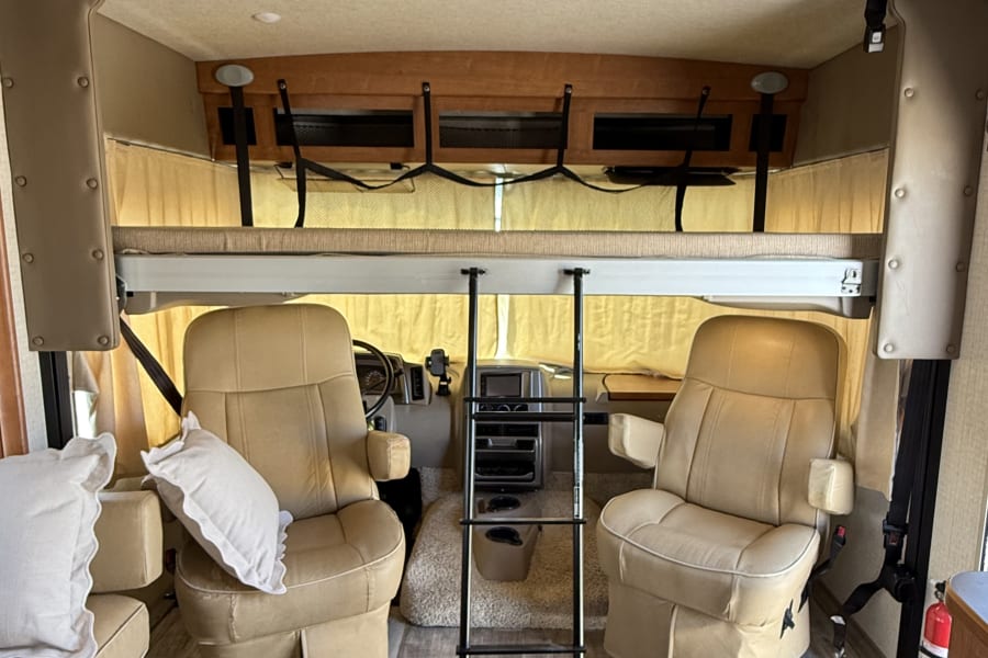 RV rental interior features a front cockpit with two plush seats, an overhead bunk, ladder, and stylish wood-look flooring.