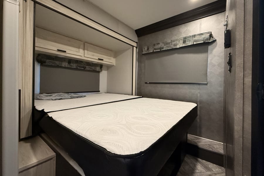 Modern RV rental bedroom featuring a clean white mattress, sleek overhead storage, and a stylish window blind.