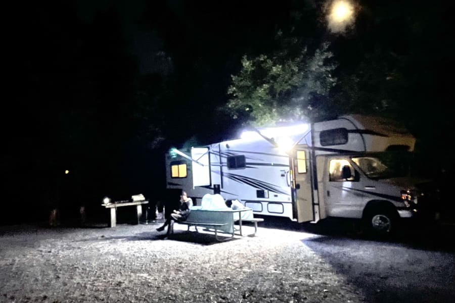 An RV rental brightly lit at a nighttime campsite, with a person sitting at a picnic table under the awning.