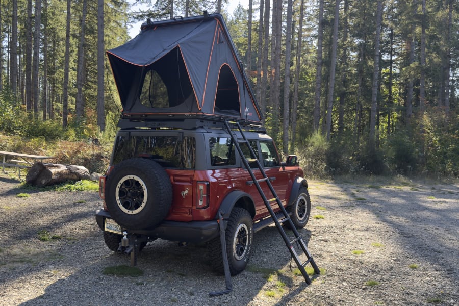 An rv rental alternative: Red Ford Bronco with rooftop tent set up for camping in a scenic forest clearing.