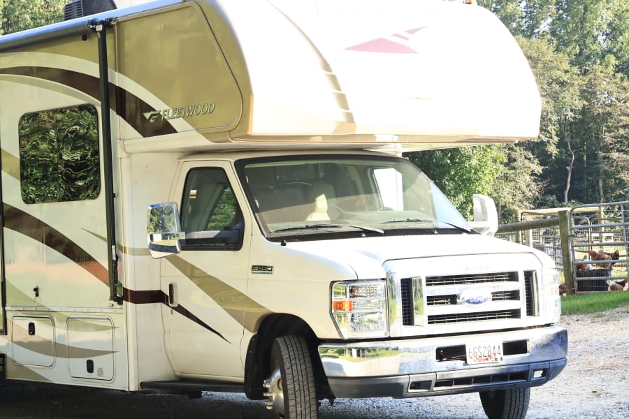 White & tan Fleetwood Class C RV rental is parked on a gravel drive, with trees in the background. Ready for adventure.