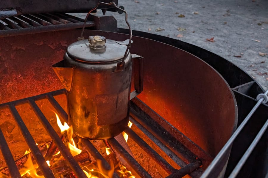 Enjoying warm campfire coffee at an RV rental site. A metal pot brews over flames, with an RV in the background.