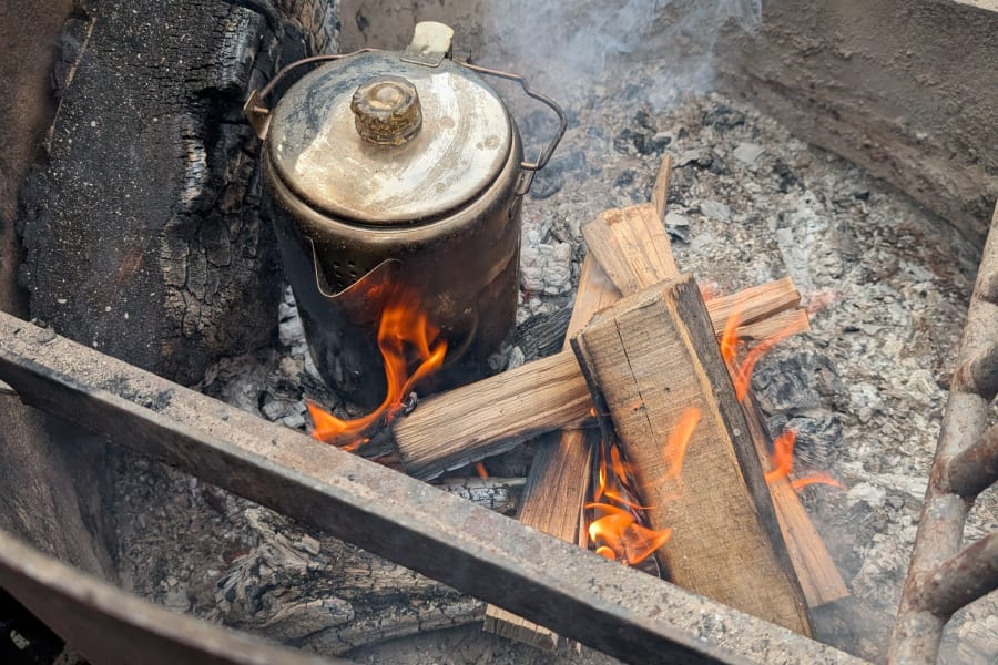 Making coffee over a campfire, a classic rv rental experience. A metal kettle sits over flames in a rustic fire pit with smoke.