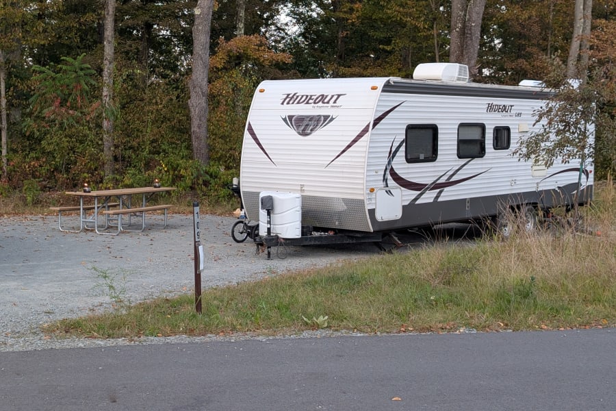 An RV rental, a white Hideout travel trailer, is parked at campsite 109 with a picnic table and trees in fall colors. Great for camping.