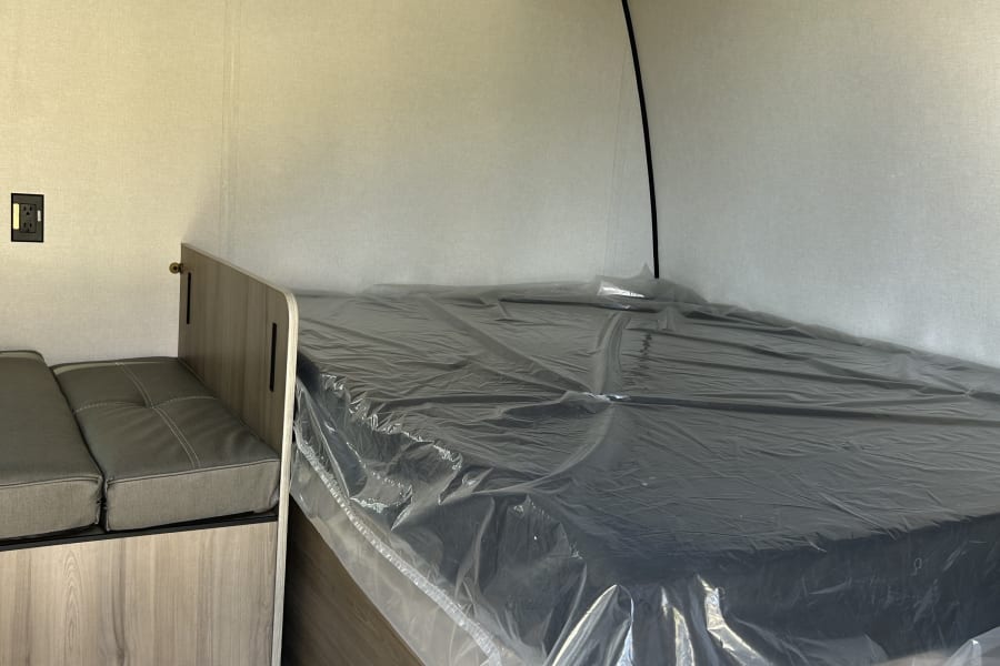 RV rental interior: new bed (plastic-wrapped), side seating, AC unit, and wall shelf in a modern travel trailer.