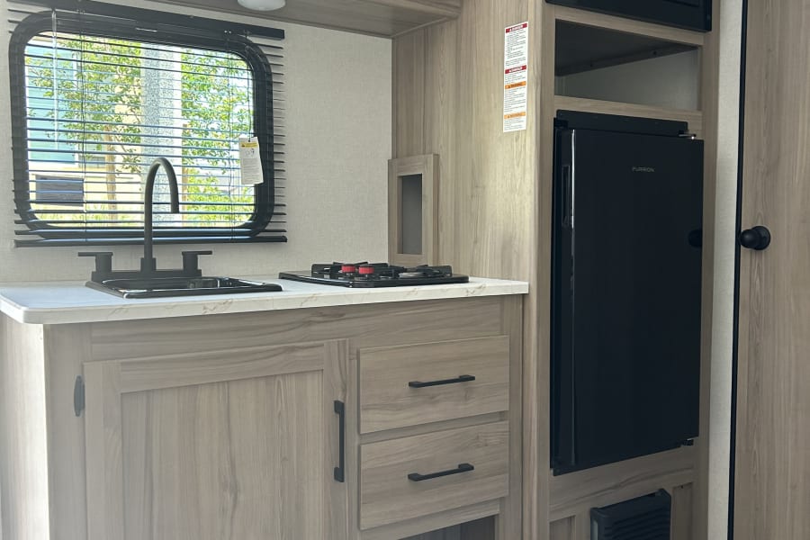 Modern rv rental kitchen featuring light wood cabinets, sink, two-burner stove, microwave, and a black compact fridge.