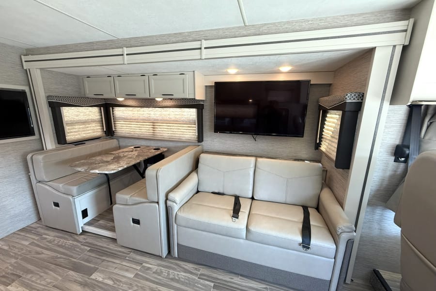Explore this modern RV rental interior featuring a dining booth, sofa with seatbelts, wall-mounted TV, and overhead cabinets.