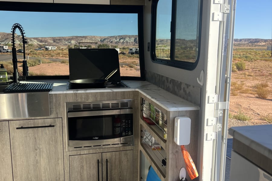 Inside an RV rental kitchen: sink, stove, microwave, dog harness, and an open door revealing a scenic desert landscape.