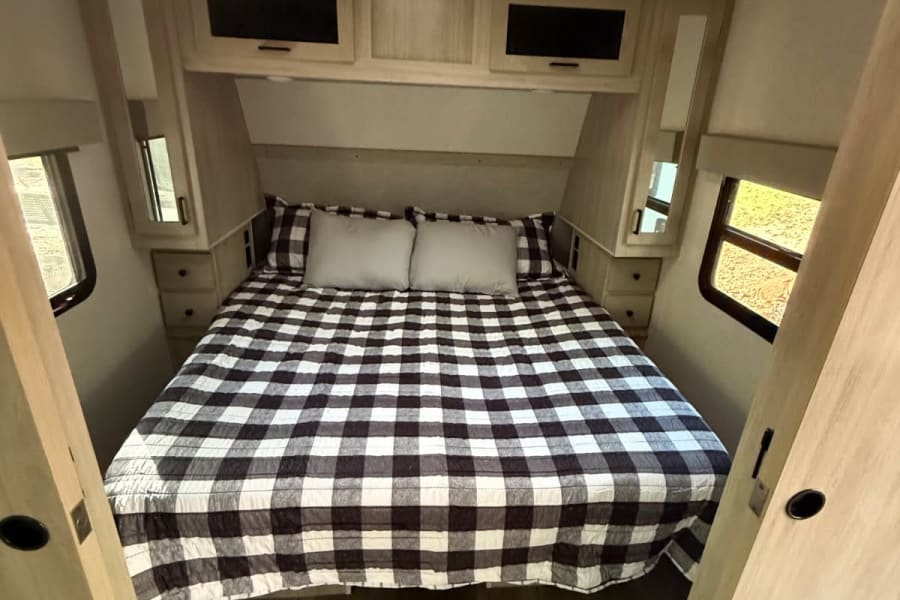 RV rental bedroom featuring a comfortable plaid bed, light wood storage cabinets, and side windows in a travel trailer.