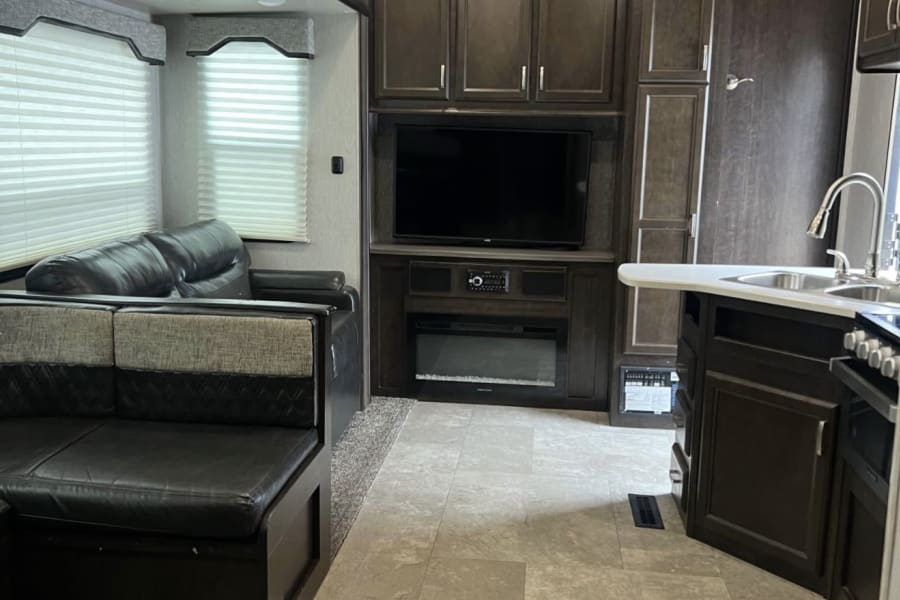 Spacious rv rental interior showing a living area with dark sofa, entertainment center, fireplace, and a kitchen on the right.