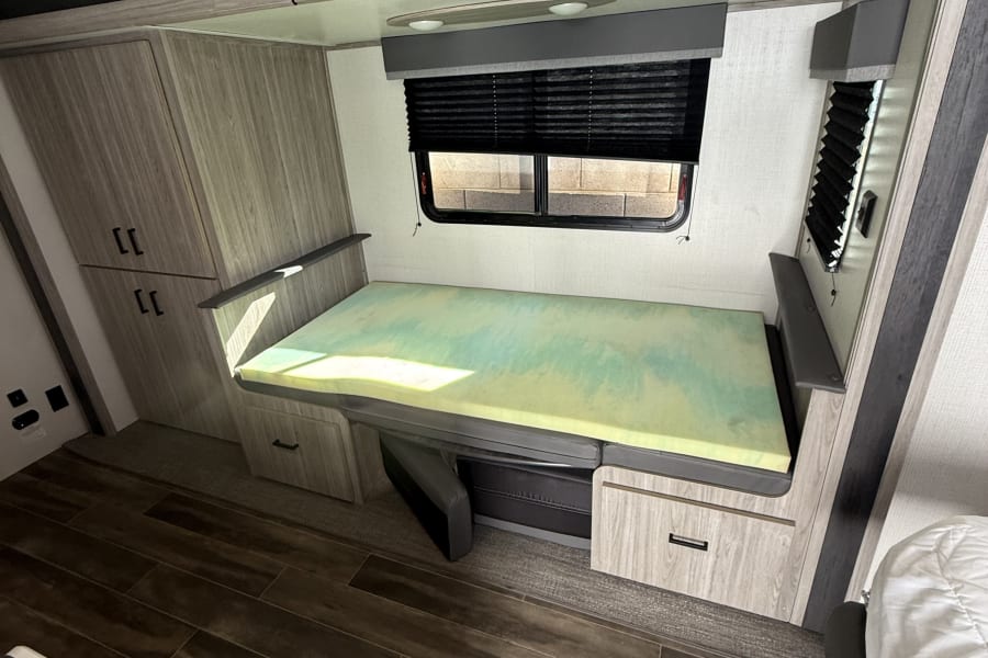 This RV rental interior features a convertible dinette bed with a green foam mattress, wood-look cabinets, and a window.