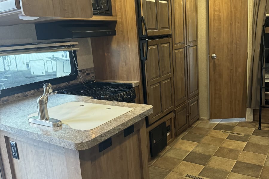 RV rental kitchen: Features a sink, stovetop, microwave, fridge, and ample wood cabinets over a warm-toned tiled floor.