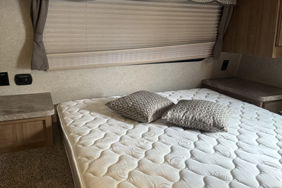 Inside an RV rental: a comfortable Serta mattress, patterned pillows, wooden cabinets, and window blinds.